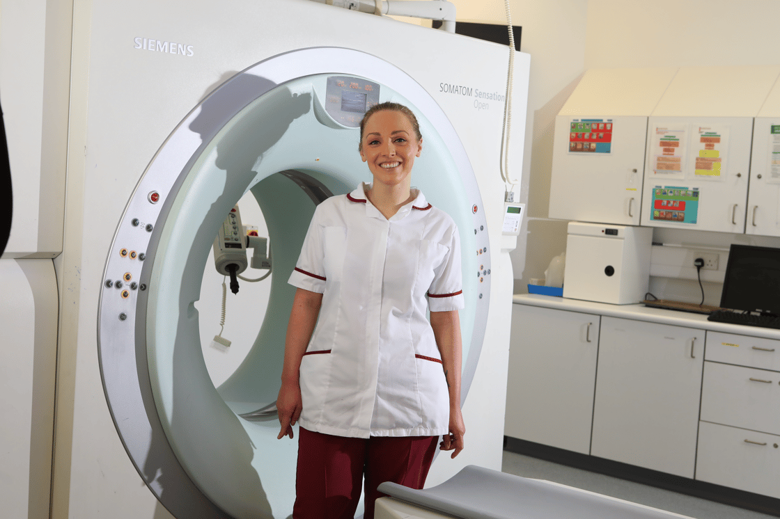 A research radiographer stands in front of a CT machine. They wear a white tunic with burgundy red piping, and red trousers
